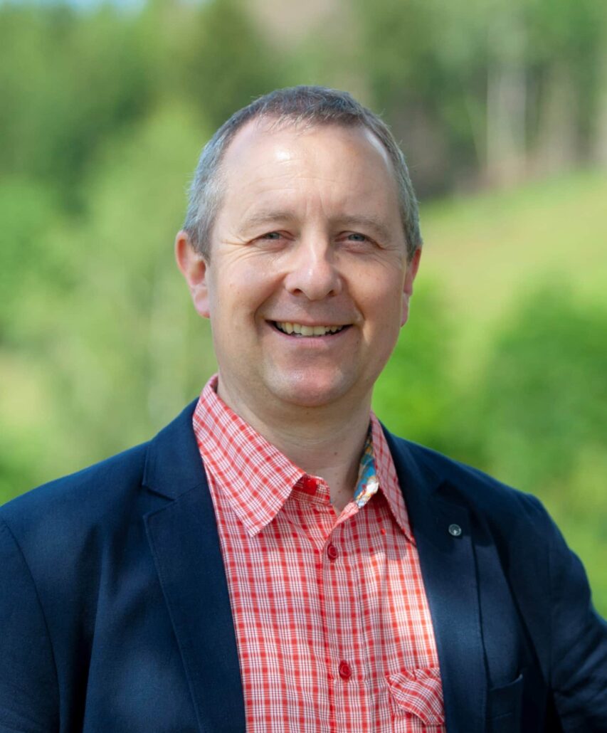 Mann mit freundlichem Lächeln in Anzug vor grüner Landschaft, repräsentiert die Werte von Unsere Grüne Glasfaser.