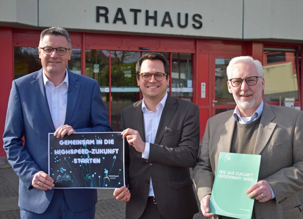 Drei Männer vor dem Rathaus präsentieren Plakate zum Start des Highspeed-Glasfaserprojekts in der StädteRegion Aachen.