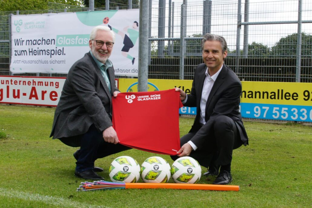 Zwei Männer präsentieren ein rotes Trikot von Unsere Grüne Glasfaser auf einem Fußballplatz mit Bällen im Vordergrund.