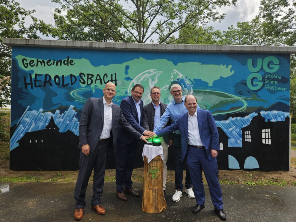 Eröffnung des Glasfaserprojekts in Heroldsbach mit Vertretern von Unsere Grüne Glasfaser vor einem bunten Wandgemälde.