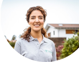 Mitarbeiterin von Unsere Grüne Glasfaser steht lächelnd vor einem modernen Wohnhaus im Freien.