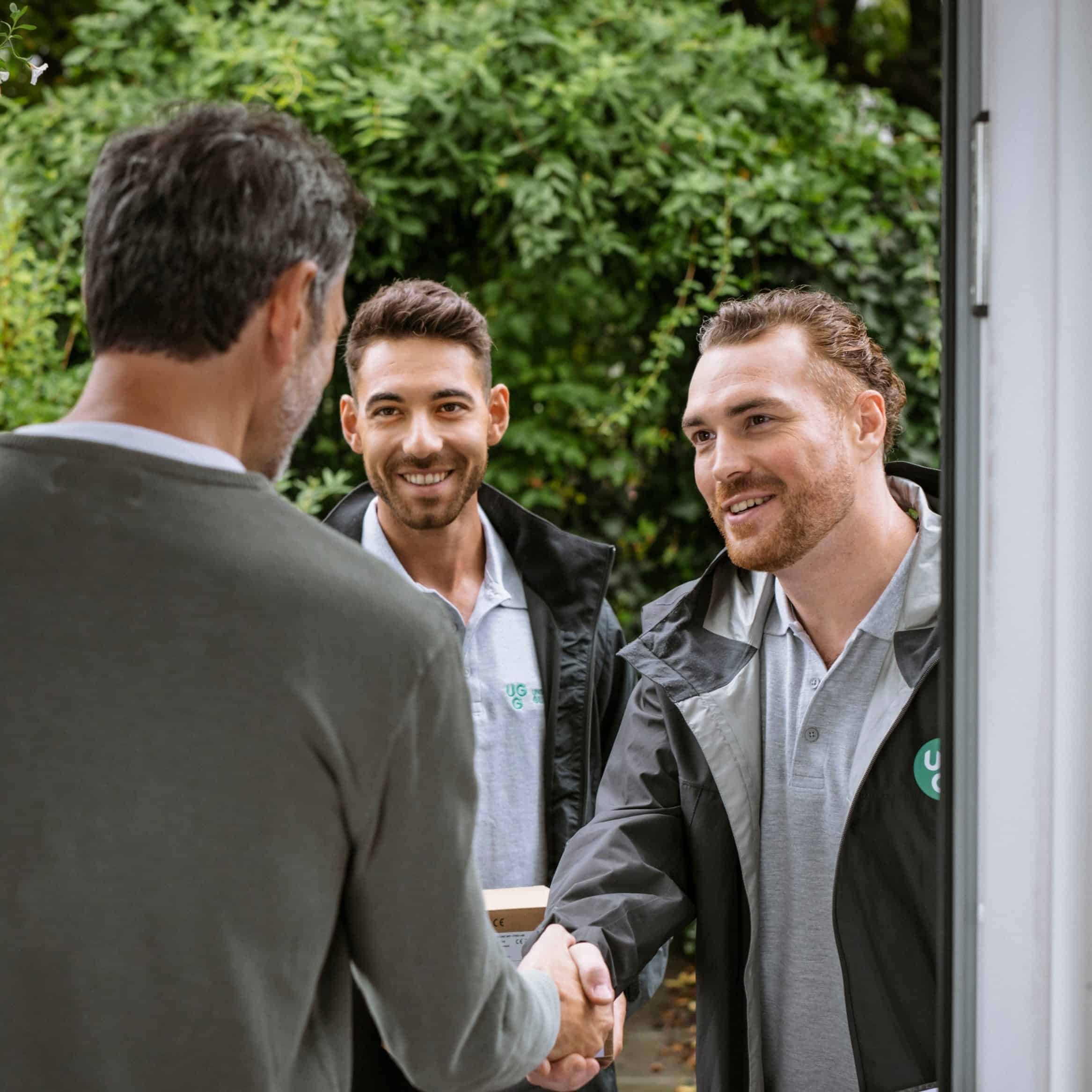 Zwei Techniker von Unsere Grüne Glasfaser begrüßen einen Kunden freundlich an der Haustür und bieten Unterstützung an.