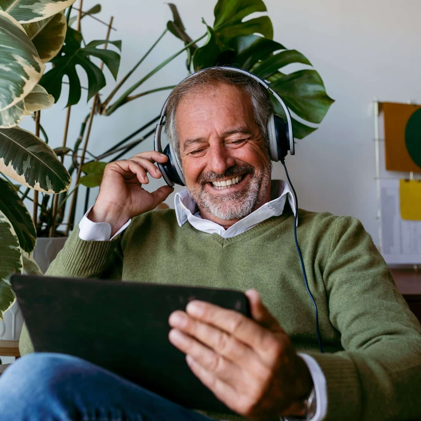 Mann mit Kopfhörern und Tablet, der entspannt in einem grünen Raum mit Pflanzen sitzt und sich digital vernetzt.