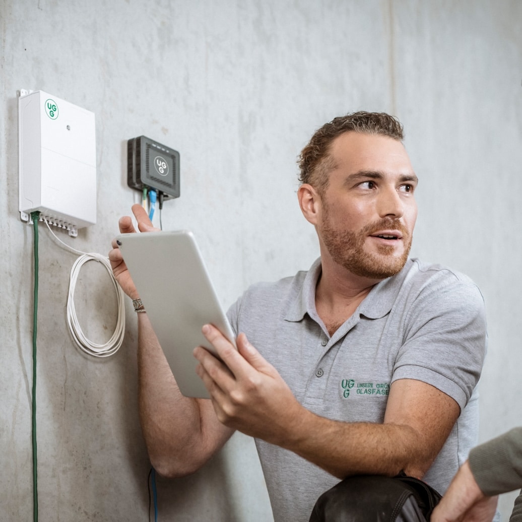 Techniker von Unsere Grüne Glasfaser installiert moderne Glasfasertechnik und erklärt die Nutzung eines Tablets.